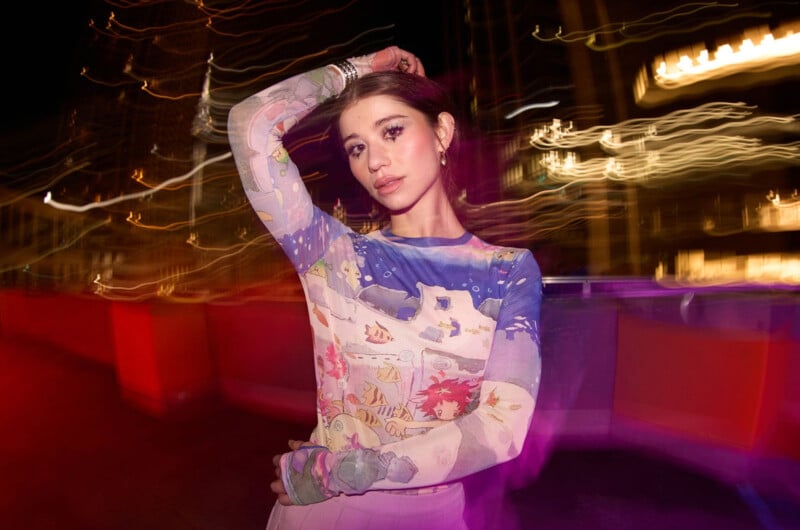 A woman in a colorful, illustrated long-sleeve top poses with one arm raised behind her head at night, surrounded by blurred, glowing city lights creating streaks and a vibrant, dynamic background.
