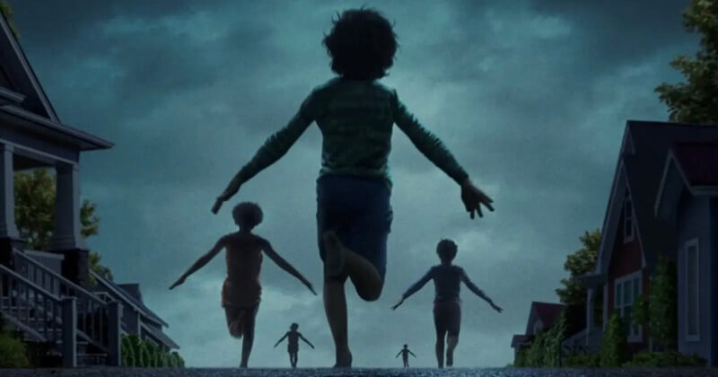 Children run down a suburban street at dusk with their arms outstretched, silhouetted against a dramatic, cloudy sky and houses lining both sides of the road.