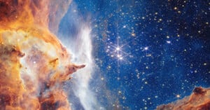 A vibrant image of outer space showing a bright star cluster surrounded by clouds of orange, yellow, and white gas and dust, with a dark blue background filled with many sparkling stars.