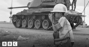 A child wearing a helmet and armband crouches and takes a photo near a large military tank on a street. A person stands atop the tank. The image is in black and white. BBC logo is in the bottom left corner.