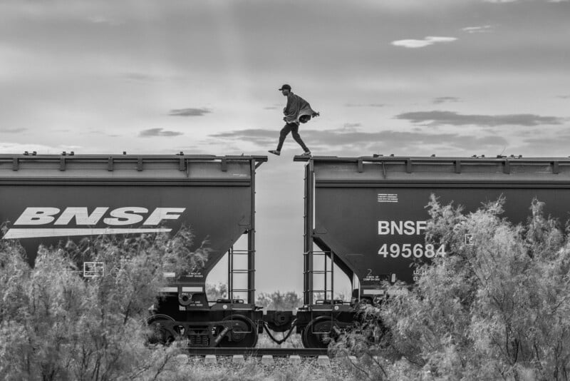 A person walks between two BNSF freight train cars, balancing on the narrow gap, with bushes in the foreground and a cloudy sky in the background. The image is in black and white.