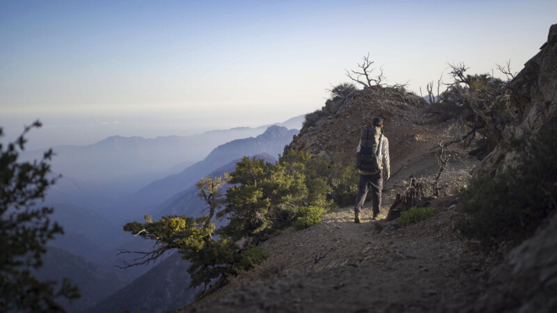 A person with a backpack hikes along a rocky mountain trail, surrounded by trees and overlooking distant misty mountains under a clear blue sky.