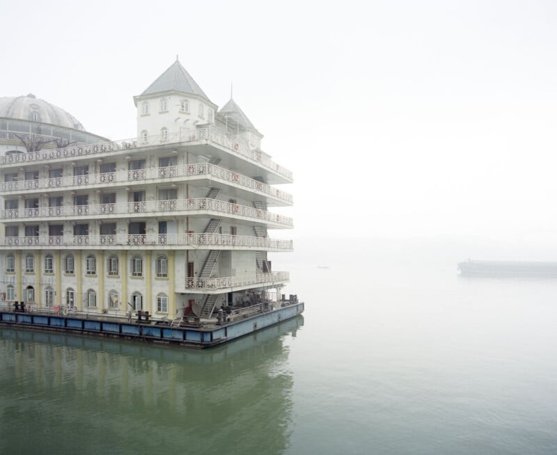 Un edificio flotante grande y ornamentado de varios pisos, como un hotel histórico, se asienta sobre aguas tranquilas y llenas de niebla, con la estructura surgiendo sobre el fondo brumoso.