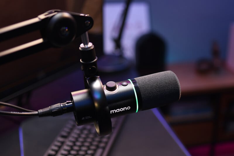A close-up of a black Maono microphone with LED lighting, mounted on a swivel arm above a computer keyboard on a desk in a softly lit room.