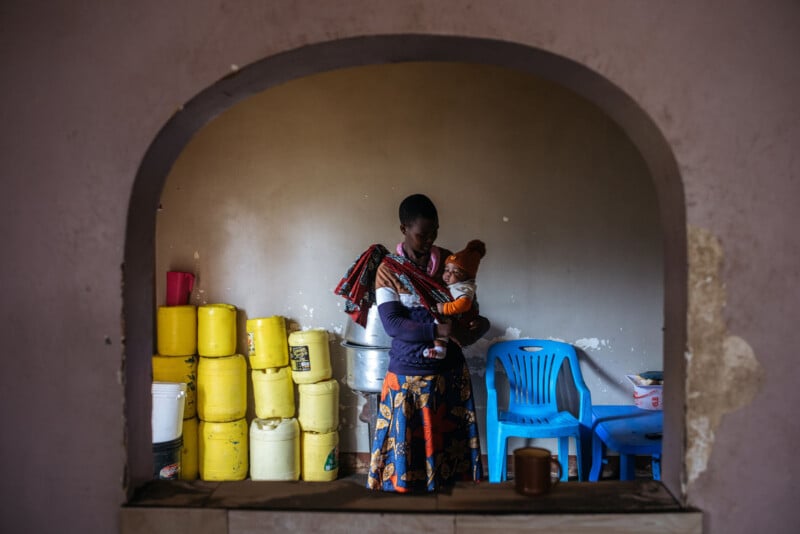 A través de la abertura arqueada se puede ver a una mujer parada en la habitación con un bebé en brazos. Detrás de ella, se apoyan en la pared recipientes de agua amarillos y sillas de plástico azules. El entorno parece simple y llano.