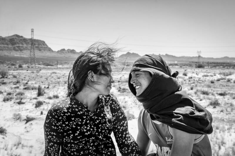 Two people smile at each other in a desert landscape, with mountains in the background. One wears a patterned shirt, the other has a shirt draped over their head. Their hair blows in the wind and both appear joyful.