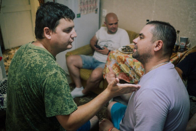 Two men are having an animated conversation in a room, with one gesturing toward the other. In the background, a third man sits on a yellow couch near a table covered with a colorful tablecloth and food items.