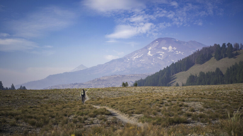 A lone hiker walks along a narrow trail through an open grassy field with a backdrop of tree-lined hills and a distant snow-capped mountain under a partly cloudy sky.