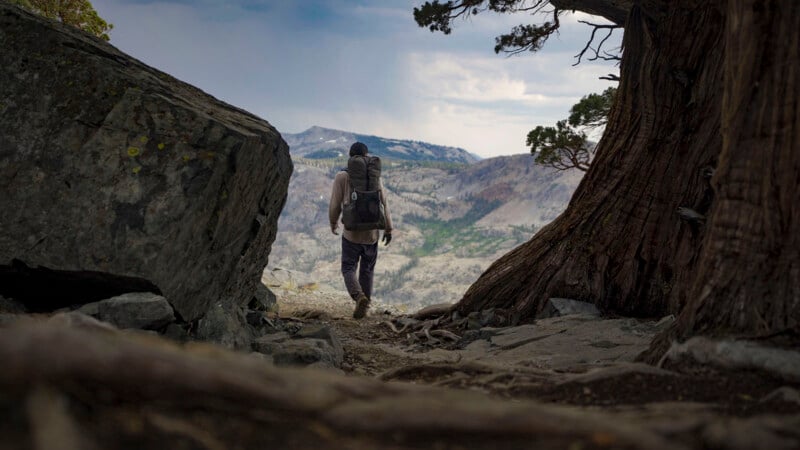 A person with a backpack hikes along a rocky mountain trail, surrounded by large trees and rocks, overlooking a scenic, rugged landscape under a cloudy sky.