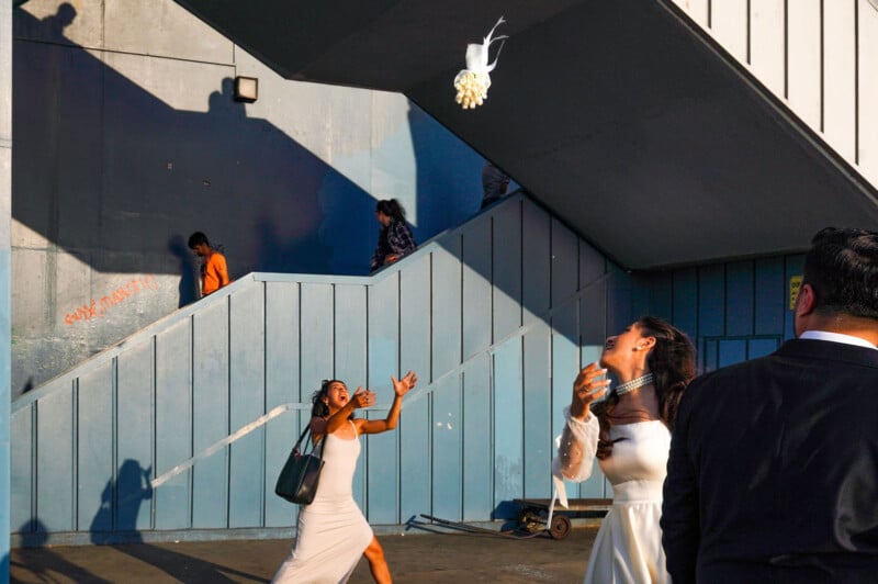 A bride throws her bouquet in the air while a woman in a white dress excitedly reaches to catch it, as other people walk by a blue staircase in the background.