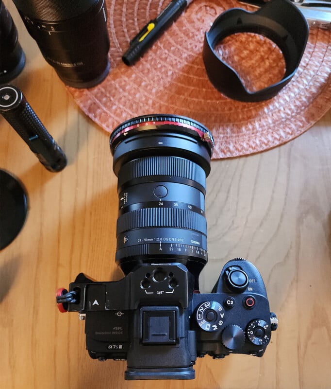A black Sony Alpha 7 III camera with a large Sigma lens attached is placed on a wooden table, surrounded by camera accessories including a lens hood and other camera equipment.