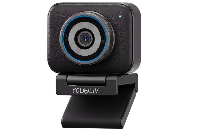 Cámara web negra rectangular compacta con una lente sobresaliente en el centro, detalle de anillo azul y una pequeña luz indicadora en el frente, montada en un soporte ajustable con la etiqueta "Yolorif.