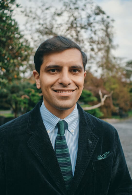 A man wearing a dark corduroy jacket, blue striped shirt, and green striped tie stands outdoors, smiling at the camera. Trees and greenery are visible in the background.