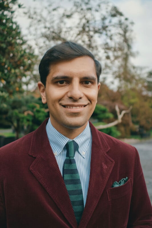 A man wearing a maroon blazer, blue striped shirt, and green striped tie stands outdoors, smiling at the camera. Trees and greenery are visible in the background.