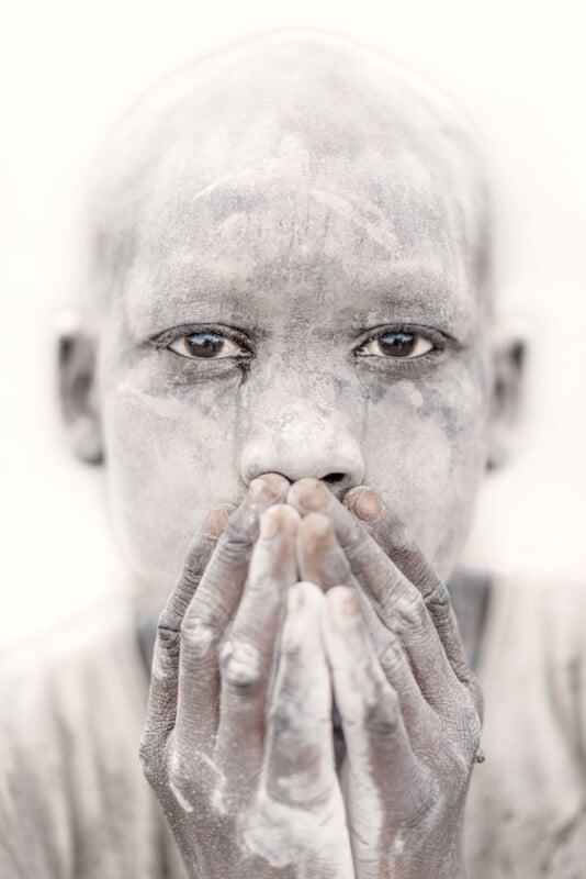 A person covered in white powder holds both hands over their mouth, gazing intently at the camera with expressive eyes. The background is bright and minimal, emphasizing the subject's face and hands.