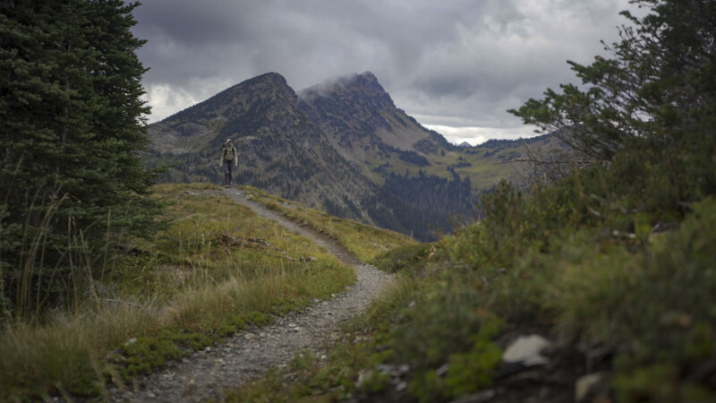 A hiker with a backpack walks along a winding mountain trail surrounded by grass and trees, with rugged mountains and cloudy skies in the background.