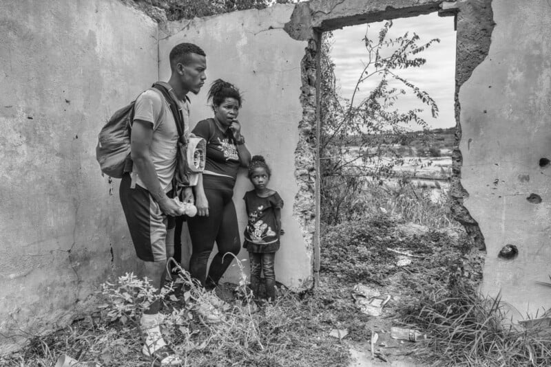 A black and white photo of a man, woman, and young girl standing in a dilapidated, roofless building, gazing through a doorway at an overgrown landscape outside. The mood appears somber and reflective.
