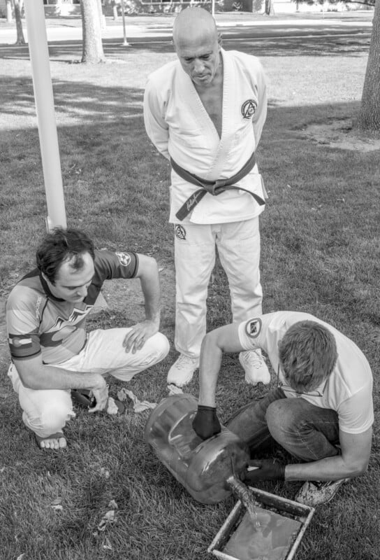 Three men outdoors examine a muddy object on the ground; one man kneels and works with gloves, another crouches beside him, while a third stands behind them in a martial arts uniform, observing.