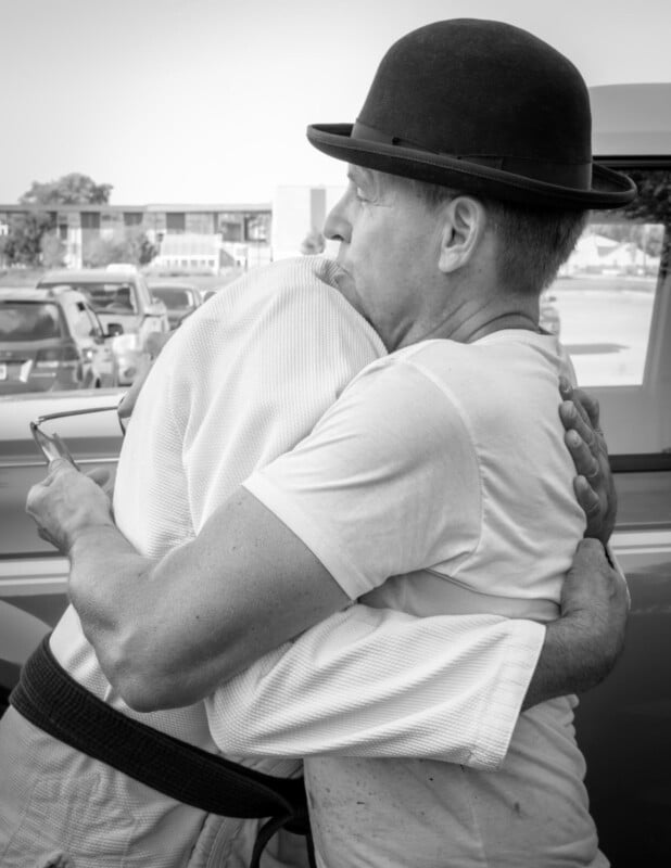 Two people embrace in a warm hug. One wears a bowler hat and a light shirt, while the other, whose face is hidden, wears a martial arts uniform and holds a phone. The scene appears to take place outdoors near parked cars.
