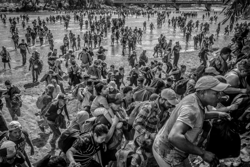 A large group of people, including families with children, cross a shallow river and climb up a muddy bank, carrying belongings. The scene is crowded and appears urgent, with many people moving together.