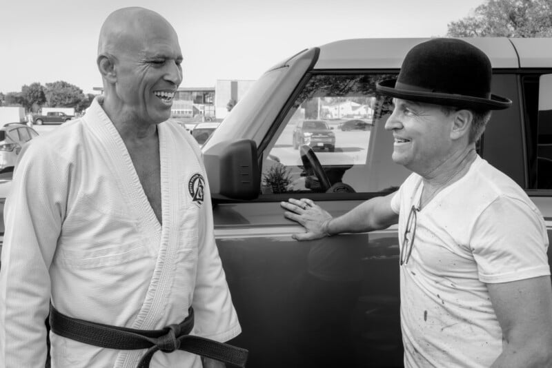 Two men stand beside a car, smiling and talking. One wears a martial arts gi with a black belt, while the other wears a bowler hat and casual clothes. The image is black and white, with parked cars in the background.