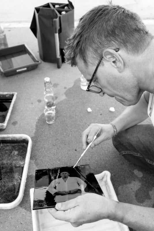 A person wearing glasses uses a brush to work on a black-and-white photograph outdoors, surrounded by trays, bottles, and photo development supplies on the ground.