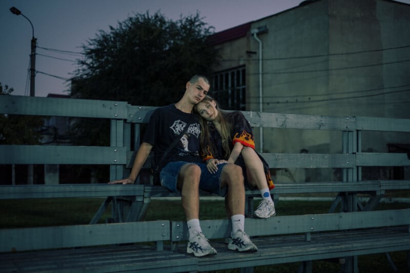 A young couple sits closely together on empty bleachers at dusk. The girl rests her head on the boy’s shoulder, and they both look relaxed. The surroundings include grass and a large building in the background.