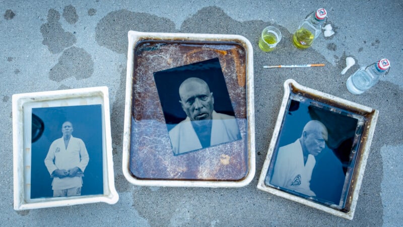 Three black-and-white photos in trays of liquid are being developed on a concrete surface; two glass bottles, a paintbrush, and a small cup are beside the trays. The photos depict a bald man in a martial arts uniform.