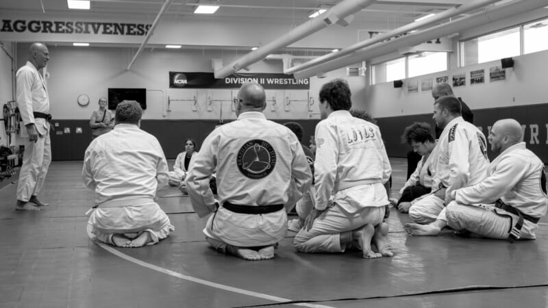 A group of people in martial arts uniforms sit on the mats, attentively listening to an instructor standing in front of them in a gym with wrestling mats and motivational banners. Black and white photo.