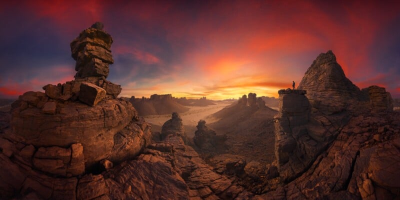 El atardecer revela un espectacular paisaje rocoso de acantilados irregulares y formaciones bajo un vibrante cielo de color naranja y violeta. Un hombre está solo en un saliente rocoso, admirando el vasto y accidentado terreno.