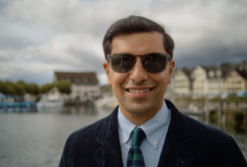 A man wearing sunglasses, a striped tie, and a dark jacket smiles at the camera. He stands outdoors near a body of water, with boats and buildings blurred in the background under a cloudy sky.