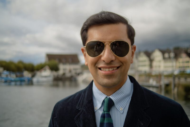 A man wearing sunglasses, a blue striped shirt, tie, and dark blazer stands smiling by a waterfront, with boats and European-style buildings visible in the blurred background.