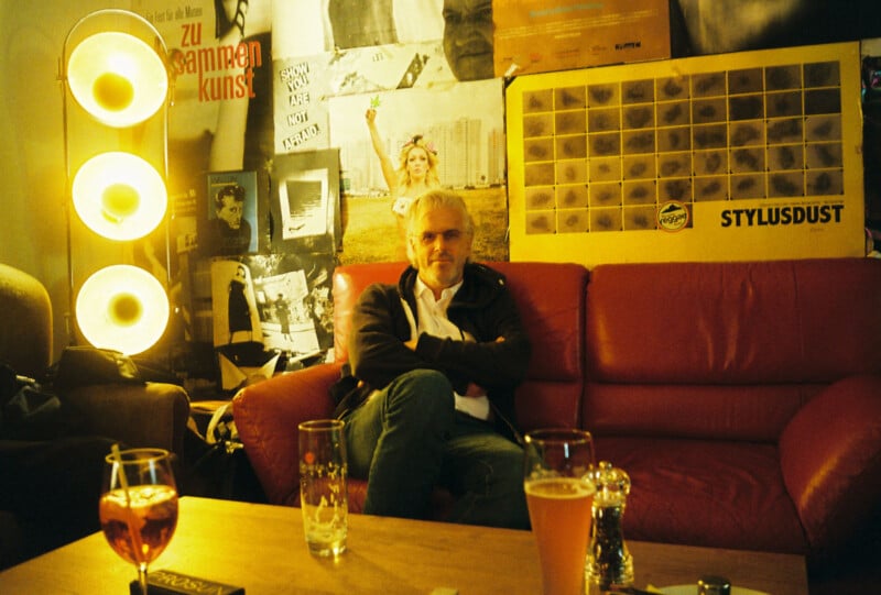 A man with gray hair sits on a red couch in a dimly lit room with posters on the wall. Three round lights glowing to the left, and various drinks and condiments are on a wooden table in the foreground.