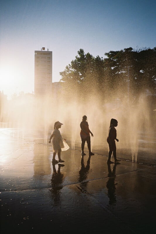 Three children walk through water fountains on a sunlit pavement, with mist rising around them. Their silhouettes are highlighted by the bright backlight. A tall building and trees appear in the background.
