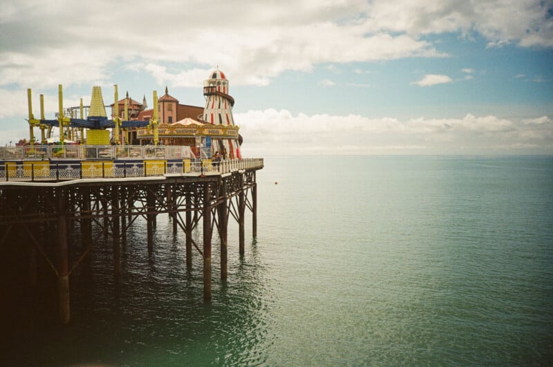 A colorful amusement park pier with a helter-skelter slide extends over calm blue-green sea under a partly cloudy sky. The horizon blends softly with the ocean.