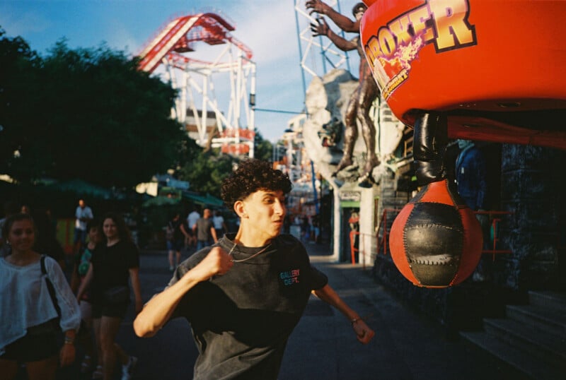 A young person punches a red boxing machine at an amusement park, with roller coasters and other visitors in the background. The sunlight highlights their focused expression and outstretched arm.