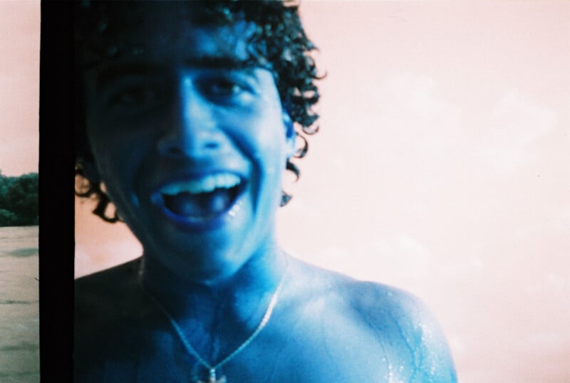 A person with curly hair, blue-tinted skin, and an open smile stands shirtless against a light background. The photo is overexposed, giving the image a surreal, playful look.