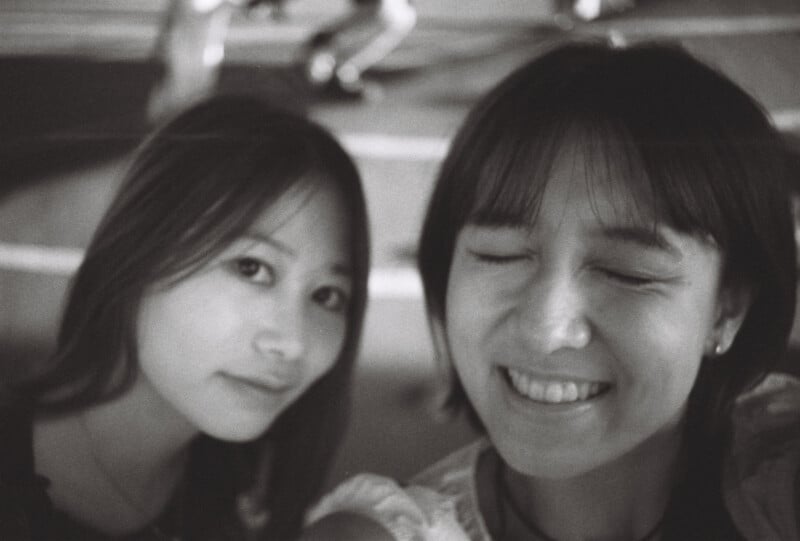 Two young women take a close-up selfie; one smiles with eyes closed while the other looks at the camera. The image is in black and white and slightly out of focus.