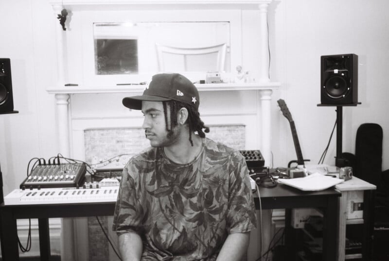A person wearing a patterned shirt and a cap sits in a music studio with keyboards, speakers, and other equipment on a desk behind them. The person is looking to the side. The image is in black and white.