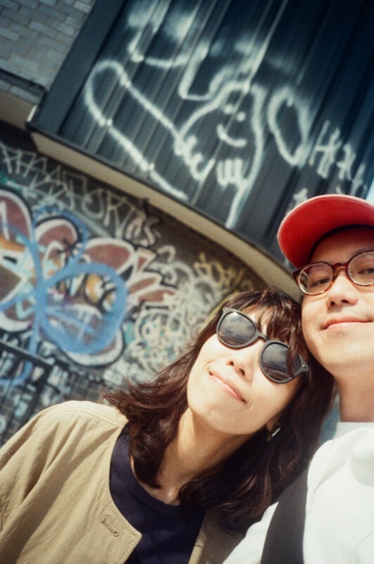 Two people wearing sunglasses smile at the camera in front of a wall covered with colorful graffiti and street art. One person wears a red cap, and the other has shoulder-length hair and a tan jacket.
