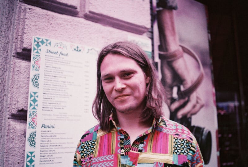 A person with long brown hair and a colorful patterned shirt stands and smiles in front of a street food menu posted on a wall. A blurred poster with a hand holding a camera is visible in the background.