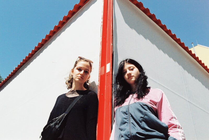 Two young women stand back-to-back against the sharp corner of a white building with a red roof, looking serious. The photo is taken from a low angle with a blue sky in the background.