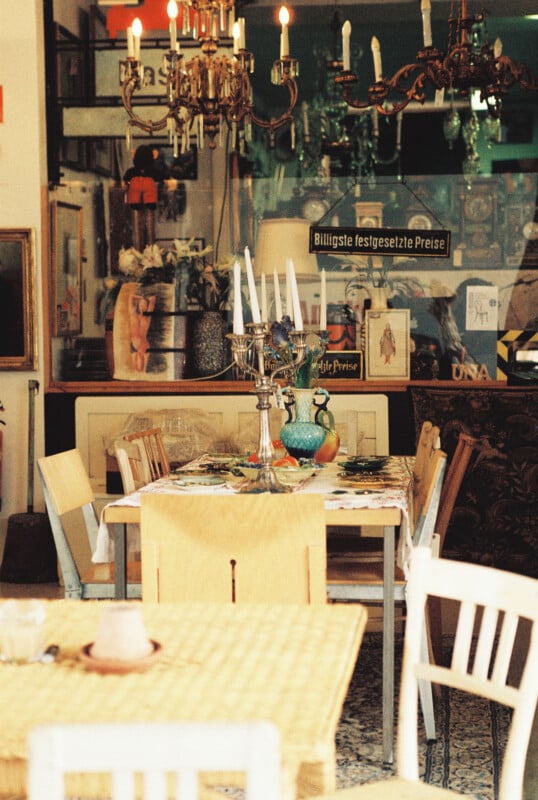 A cozy, vintage dining area with wooden tables and chairs, a candelabra centerpiece, eclectic decor, chandeliers, framed art, and a German sign reading "Billigste festgesetzte Preise" in the background.