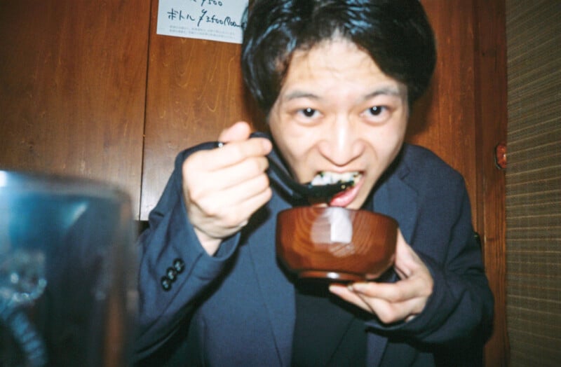 A person in a dark jacket is eating food with a spoon from a wooden bowl, looking directly at the camera in a warmly lit room with wooden walls.