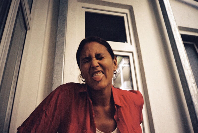 A person wearing a red shirt stands in front of a building, playfully scrunching their face and sticking out their tongue, with closed eyes and relaxed posture.