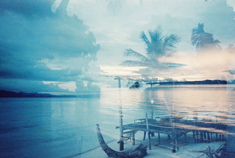 A dreamy, blue-tinted double exposure shows a calm tropical beach with palm trees, wooden lounge chairs, and clouds reflected on the water at sunset, creating a serene, surreal atmosphere.