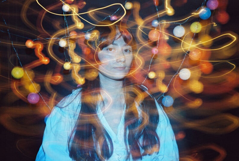 A woman with long hair and a white shirt stands in front of colorful string lights. Light trails swirl around her, creating a dreamy, abstract effect against a dark background.
