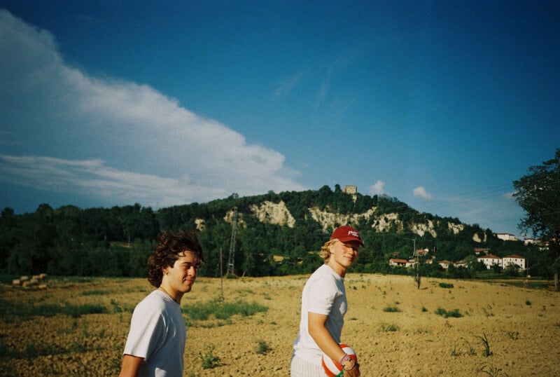 Two people in white shirts walk through a sunlit, sandy field holding a ball, with a forested hill, a castle, and houses in the background under a blue sky with clouds.