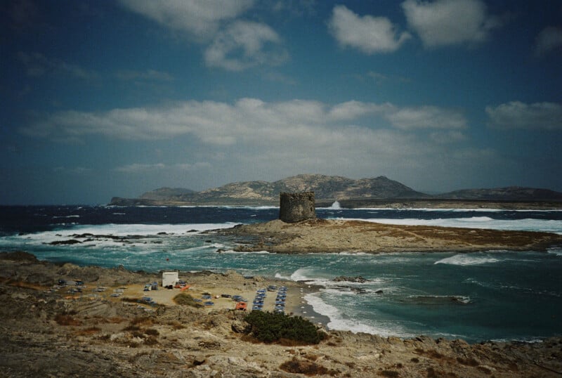 A rocky coastal landscape with a medieval stone tower on a small island, turquoise waves crashing on the shore, and distant mountains under a partly cloudy sky. Cars are parked along the sandy beach in the foreground.
