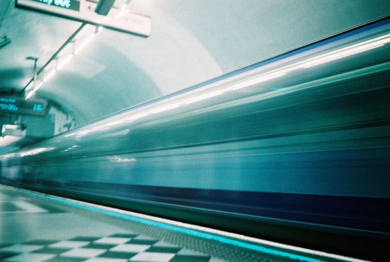 A blue and white train speeds through an underground subway station, creating a motion blur. The curved ceiling, fluorescent lights, and tiled platform are visible, along with a blurry overhead sign and geometric floor pattern.